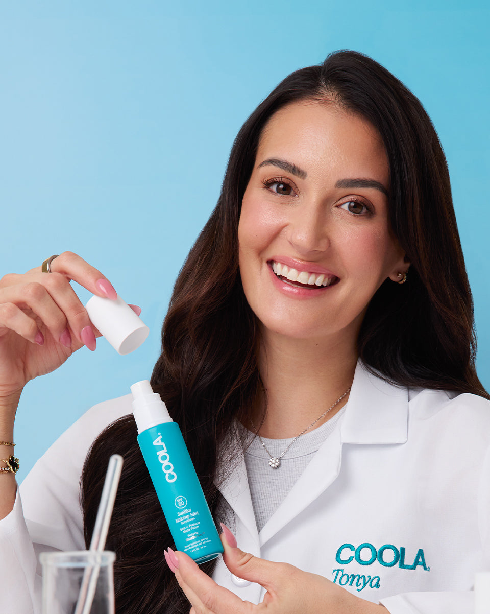 Woman holding a Coola Makeup Mist bottle smiling on a blue background