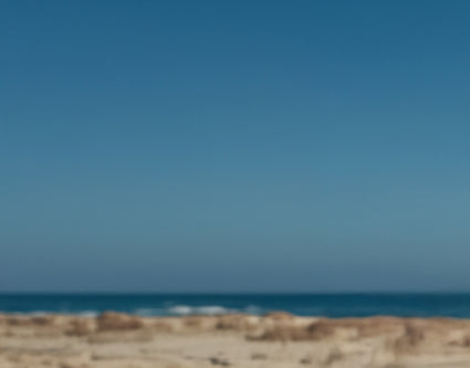 Blurred image of a beach with sand and ocean waves under a clear blue sky.
