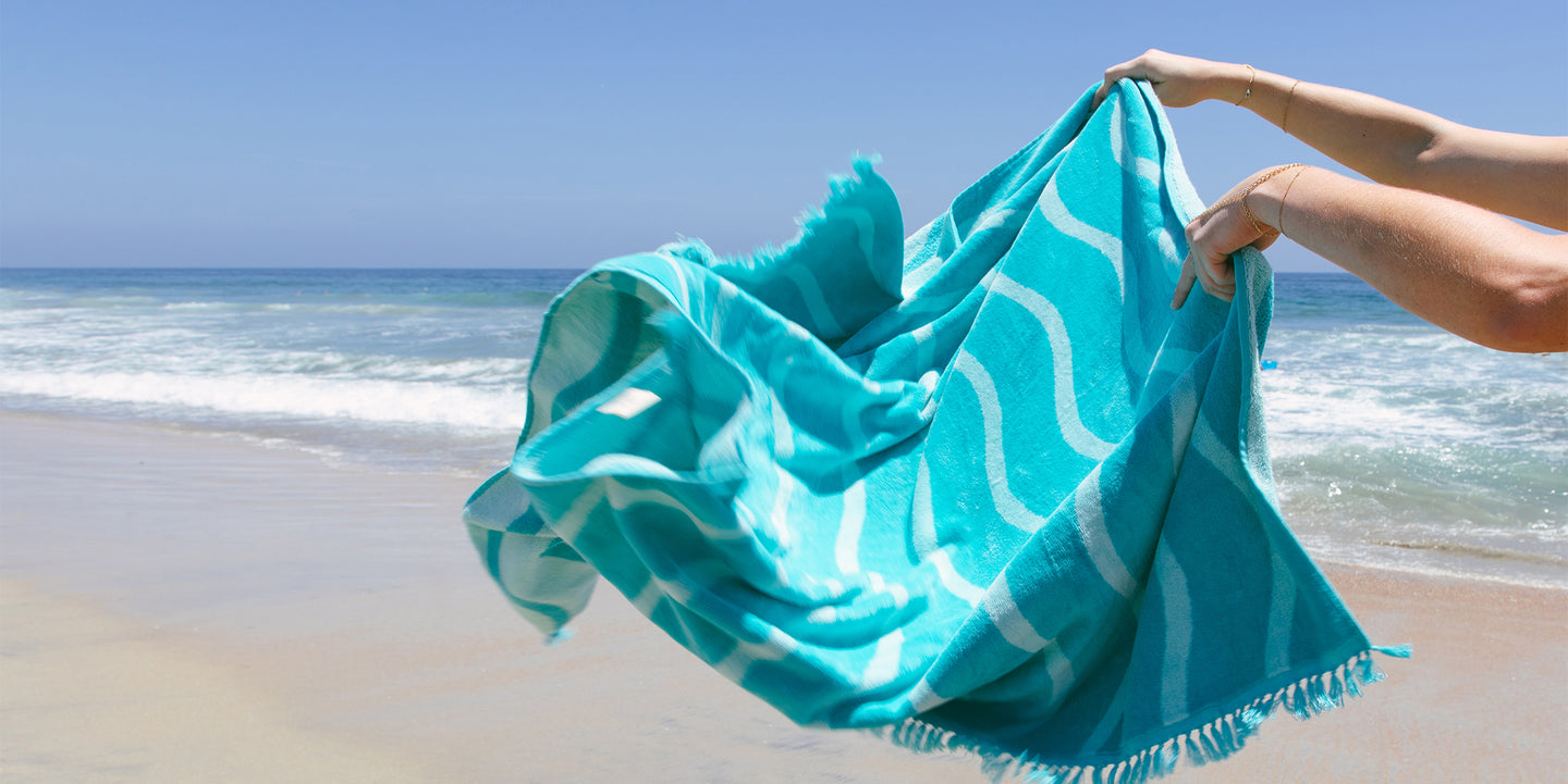 Person holding a turquoise and white patterned towel fluttering in the breeze on a sunny beach with ocean waves in the background