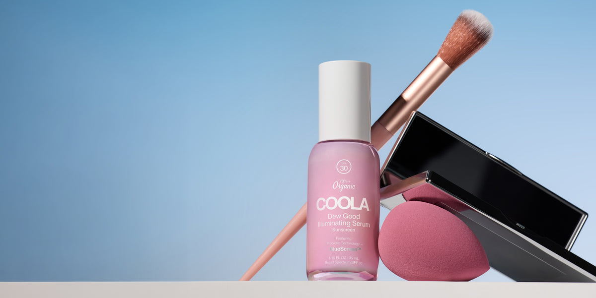 Pink COOLA skincare bottle beside a makeup brush and open black compact with pink powder against a soft blue background