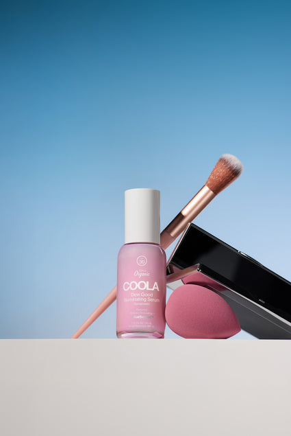 COOLA pink skincare bottle with a makeup brush and pink compact arranged against a soft blue background