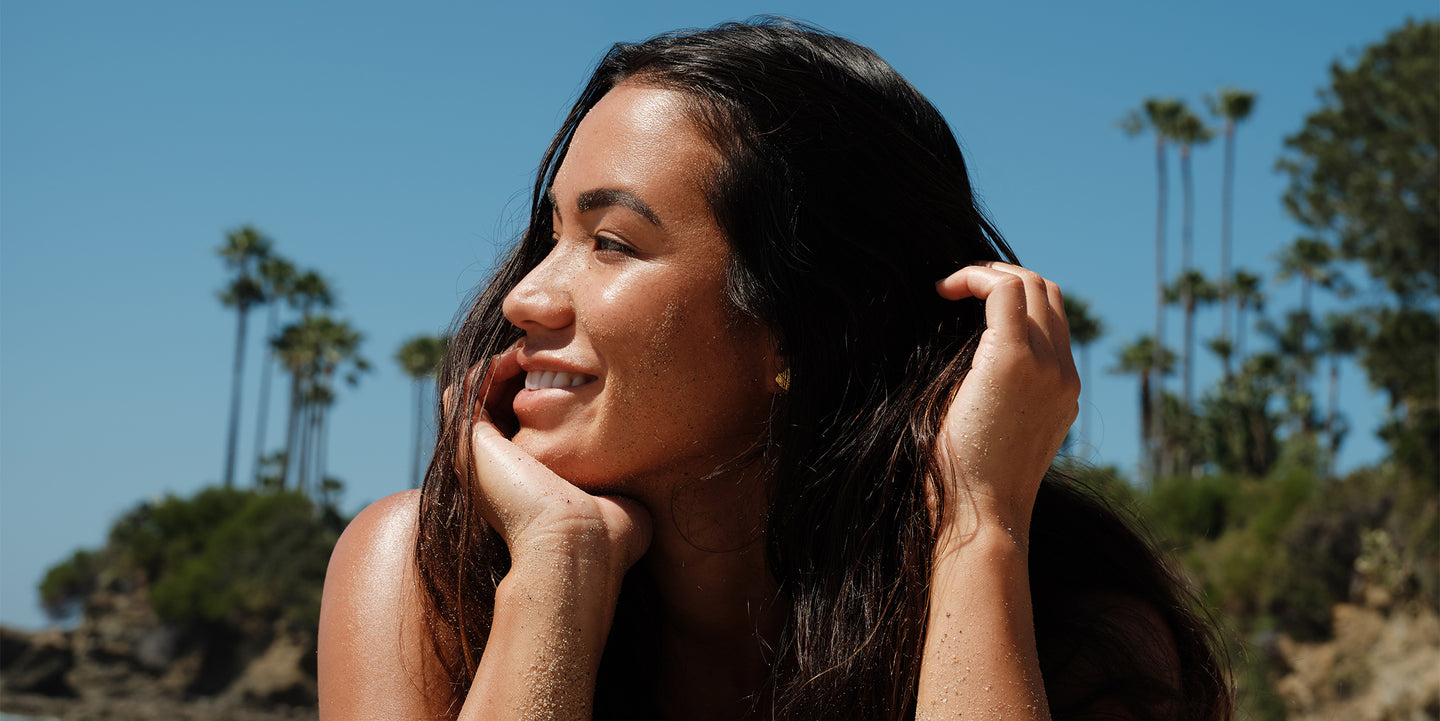 Smiling woman with long dark hair outdoors in bright sunshine surrounded by palm trees and greenery under a clear blue sky