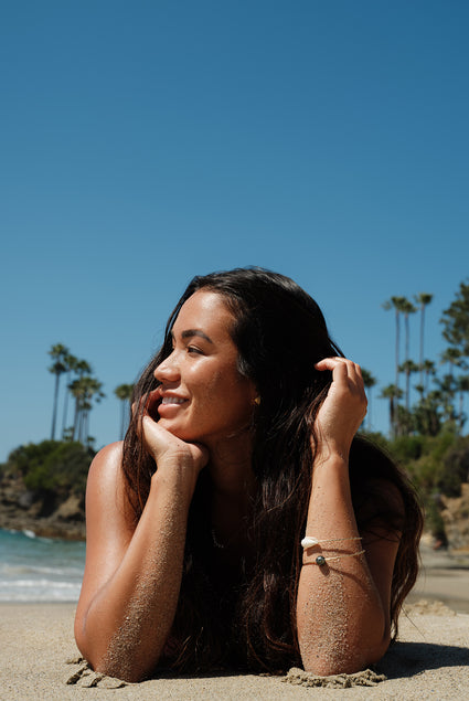 Smiling woman with long dark hair resting on a sandy beach beneath a clear blue sky with palm trees and ocean waves in the background