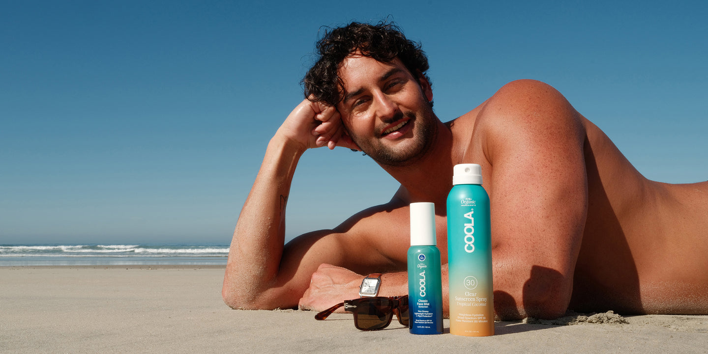 Henry laying on the beach next to sunglasses and COOLA sunscreen