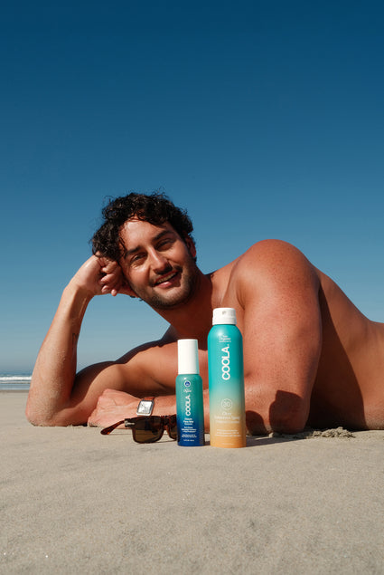 Henry laying on the beach next to sunglasses and COOLA sunscreen