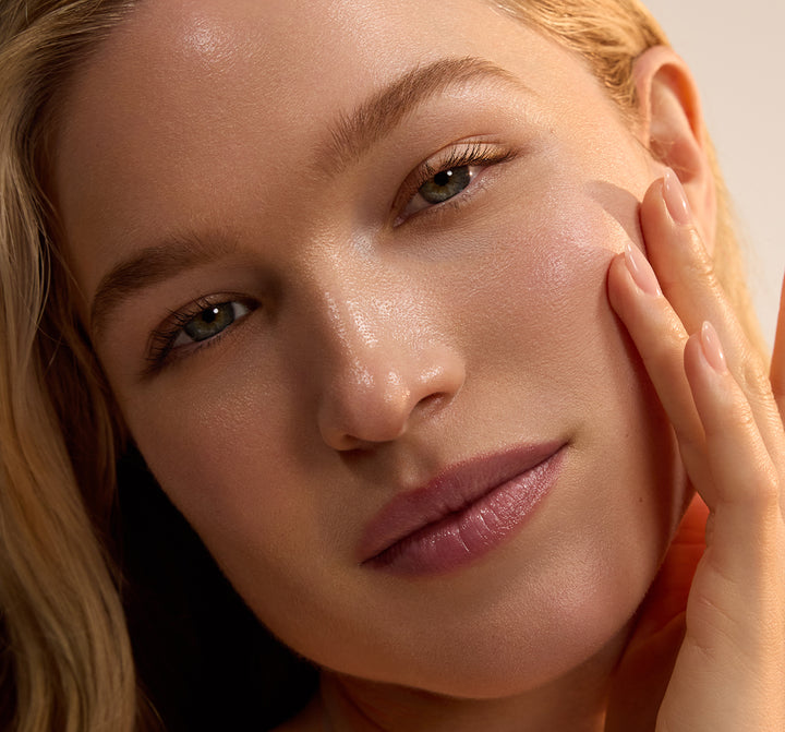 Close up portrait of a woman with dewy skin gently touching her cheek in warm natural light and looking toward the camera