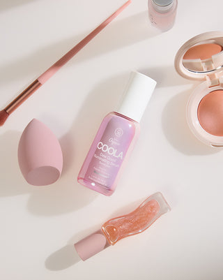 Flat lay of a pink COOLA bottle with a makeup brush sponge compact powders and a glossy tube arranged on a soft white background