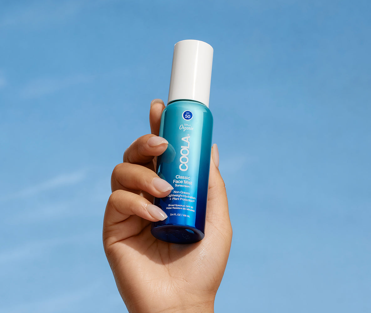 Hand holding COOLA Classic Face Mist SPF 50 bottle with blue sky background.