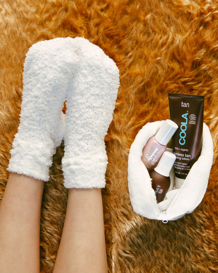 Feet wearing cozy white fuzzy socks next to a white fuzzy pouch containing COOLA tan lotion and two nail polish bottles, all placed on a soft brown fur surface.