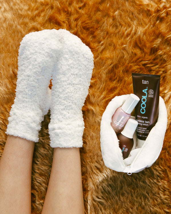 Feet wearing cozy white fuzzy socks next to a white fuzzy pouch containing COOLA tan lotion and two nail polish bottles, all placed on a soft brown fur surface.