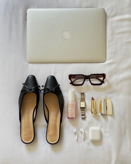 Flat lay of black ballet flats, sunglasses, gold watch, lipstick with case, Coola sunscreen bottle, wireless earbuds with case, and a closed silver laptop on a white fabric background.