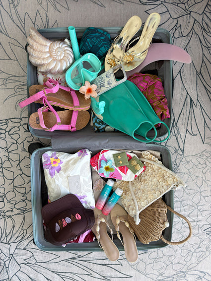 Open suitcase filled with summer essentials including pink sandals, snorkeling gear, gold slip-on shoes, tropical patterned clothes, COOLA sunscreen bottles, and beach-themed accessories on a floral patterned surface.