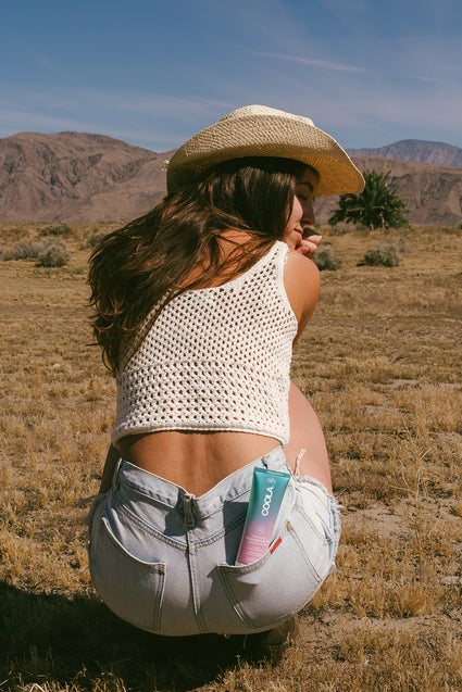 Woman wearing a straw hat and white crochet top squatting in a dry, open landscape with mountains in the background, a tube of COOLA sunscreen visible in her back pocket