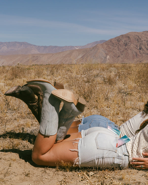 Person lying on their stomach in a dry desert landscape wearing cowboy boots, distressed denim shorts with sunscreen tubes tucked in the back pocket, and a light sweater.