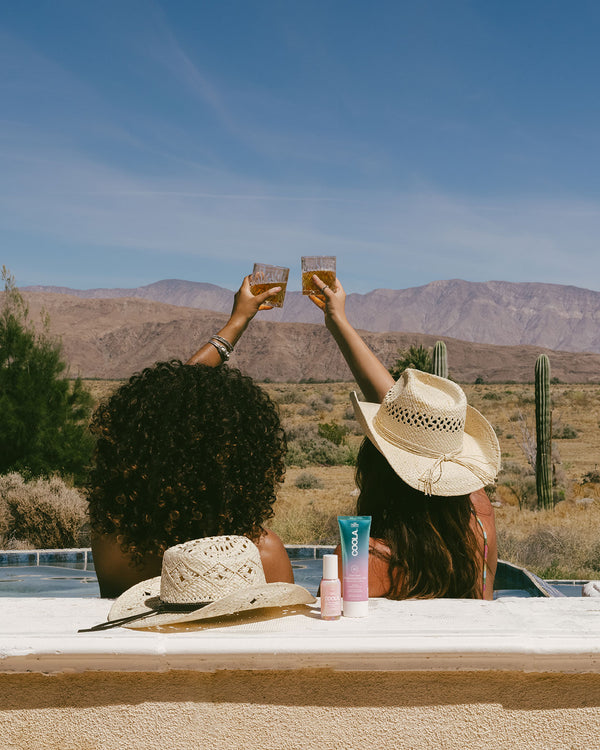 Two women in sun hats sitting by a pool in a desert landscape, raising glasses with COOLA sunscreen products and an additional hat resting nearby.