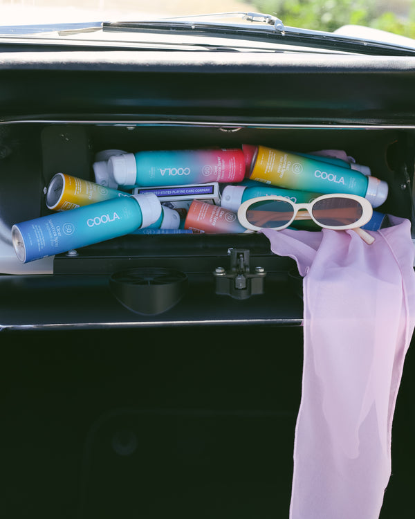 A car glove compartment filled with colorful COOLA sunscreen bottles, a pair of white-framed sunglasses, and a light pink scarf casually draped inside.