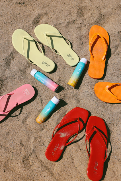Four pairs of colorful flip-flops in yellow, orange, pink, and red are arranged in a circle on sandy ground with four tubes of COOLA sunscreen placed in the center.