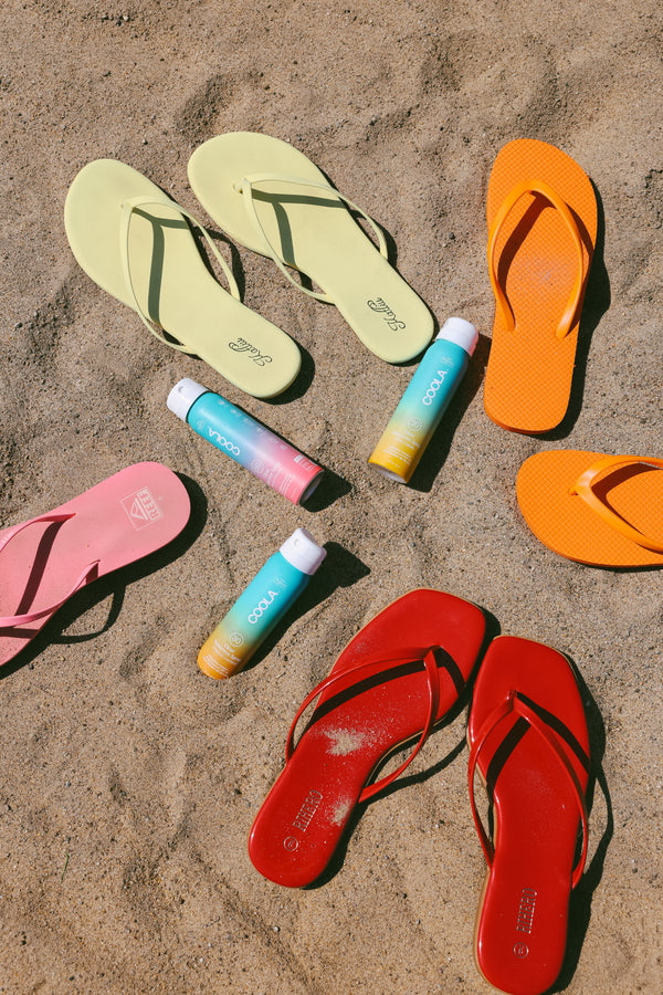 Four pairs of colorful flip-flops in yellow, orange, pink, and red are arranged in a circle on sandy ground with four tubes of COOLA sunscreen placed in the center.