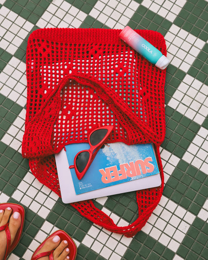 Red knitted bag on green and white tiled floor containing red sunglasses and a magazine titled "SURFER," next to a COOLA sunscreen stick and red sandals with painted toenails visible at the bottom.