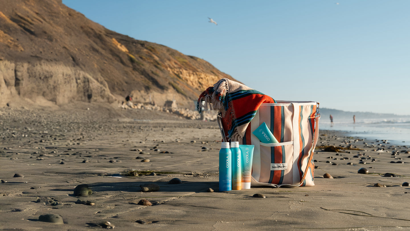 Collection of sunscreen products sitting beside patterned tote bag with scenic beach scene in the background