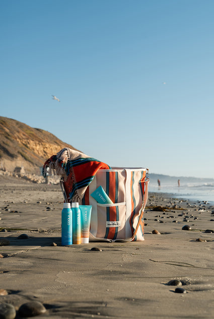 Collection of sunscreen products sitting beside patterned tote bag with scenic beach scene in the background