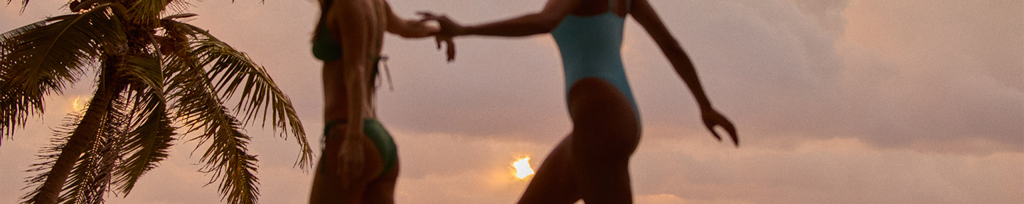 Two people in swimsuits standing on a beach with palm trees and sunset in the background
