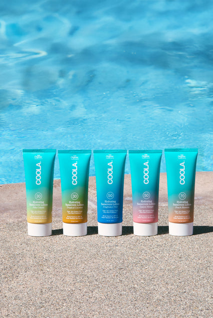 Five tubes of different Coola body lotions lined up on ground with pool in background