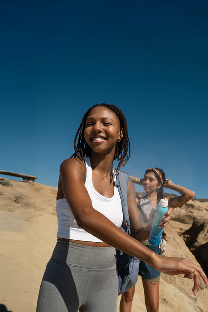 Two women in coastal setting on a hike with woman in front smiling applying Coola spray sunscreen