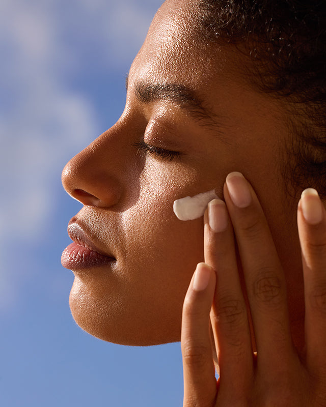 Woman applying lotion to her cheek with a blue sky background