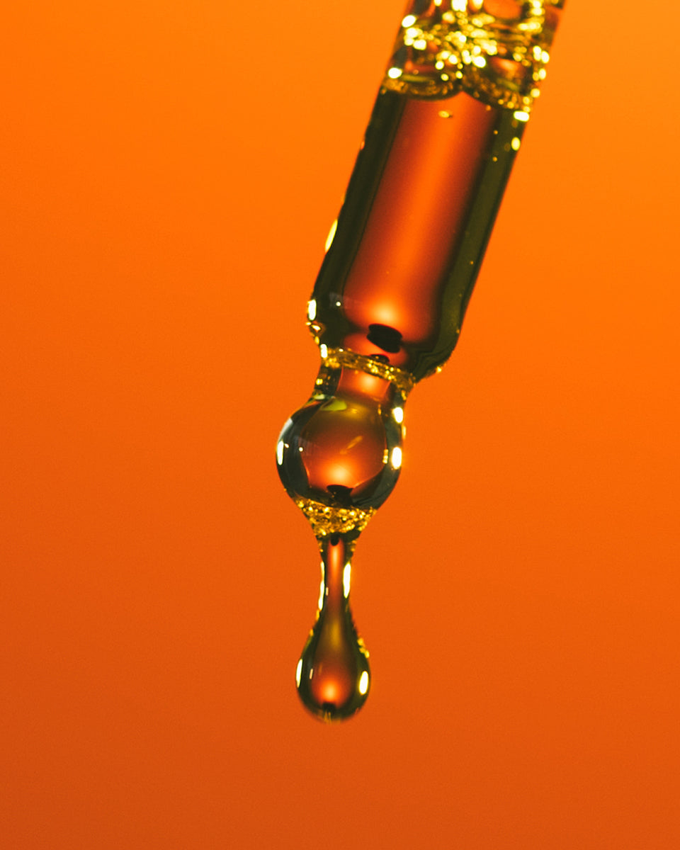 Macro image of face oil sunscreen dropper dispensing a drop of oil against a golden orange background