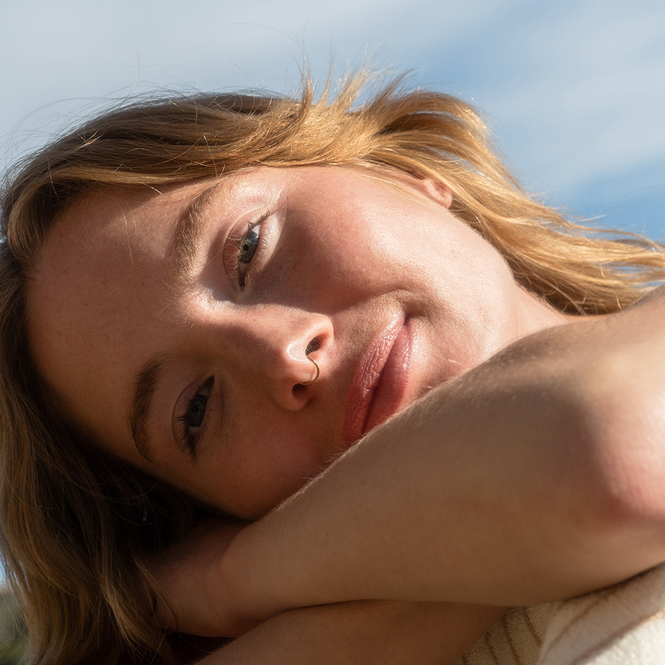 Close-up of woman resting face on arms with soft smile and blue sky background.