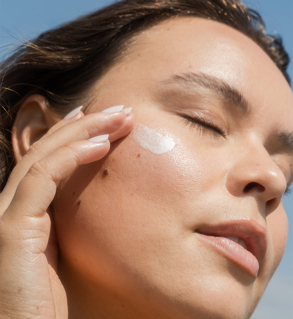 Close-up of woman applying sunscreen lotion to cheek with blue sky background