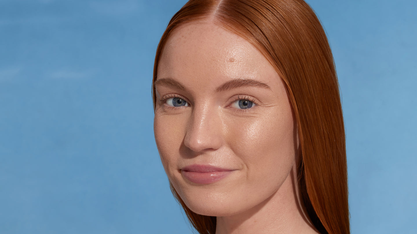 Model with long straight red hair against a blue background, with dew good applied. Skin is glowy and illuminated.