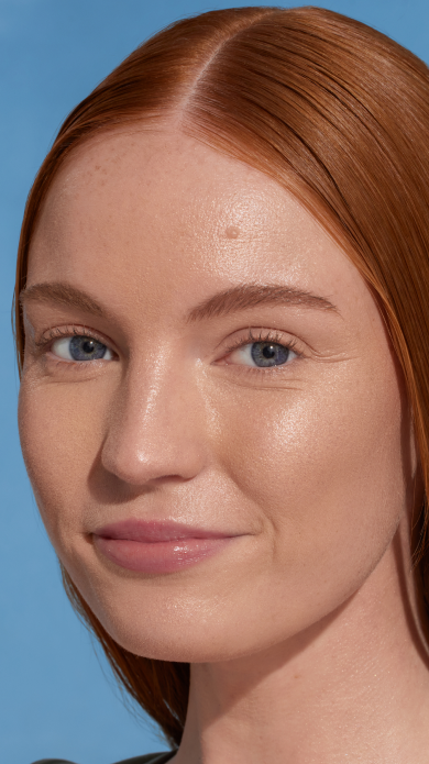 Model with long straight red hair against a blue background, with dew good applied. Skin is glowy and illuminated.