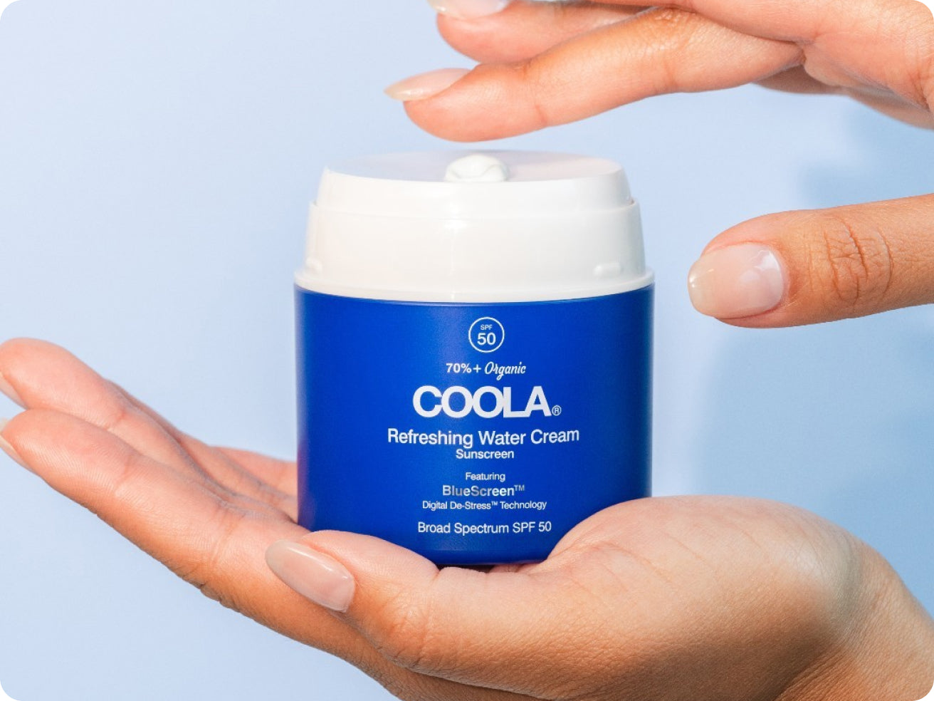 A person holds a blue container of COOLA Refreshing Water Cream sunscreen with SPF 50 against a light blue background, dispensing formula from the white cap