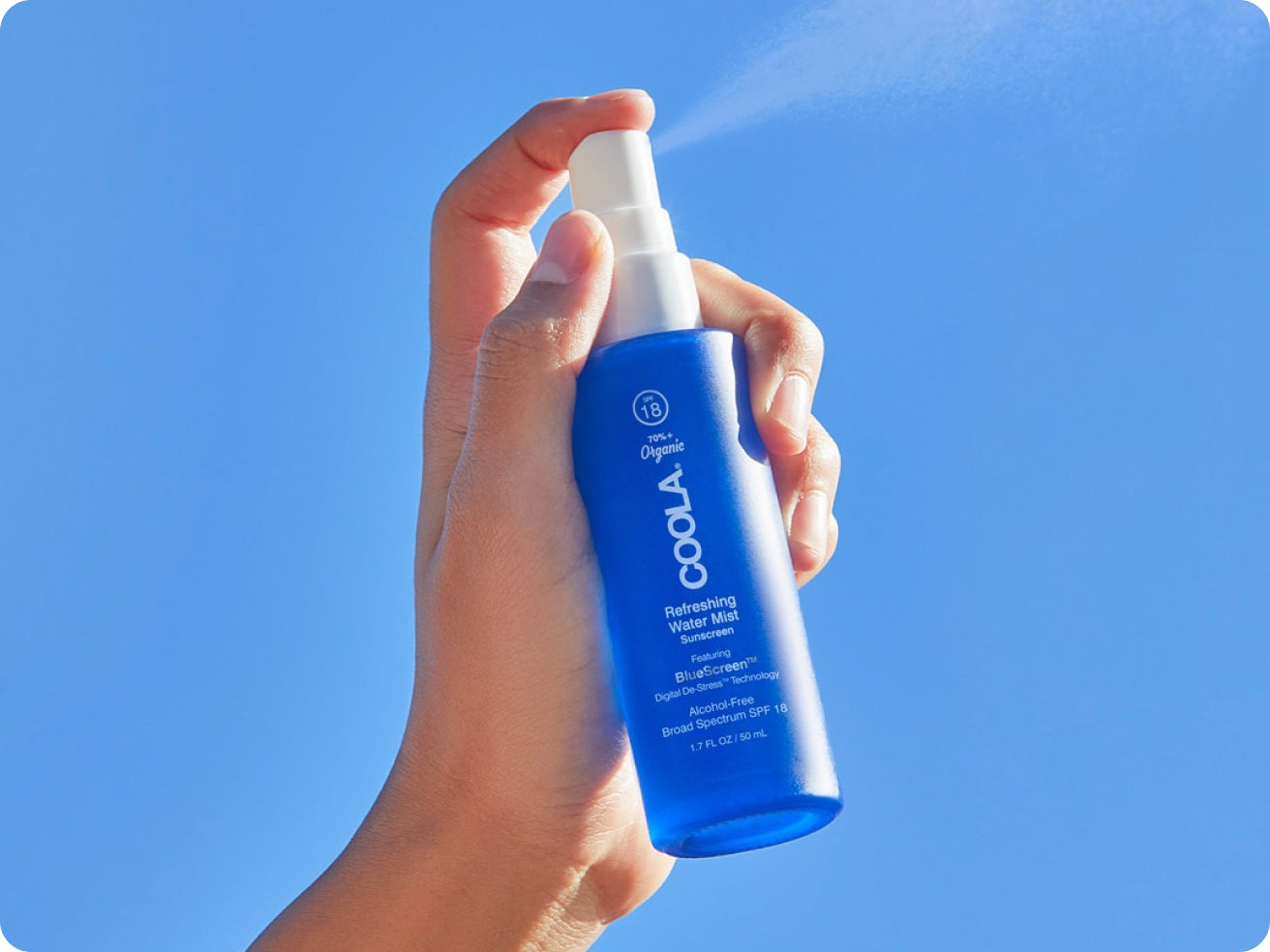 A hand holds a blue COOLA Refreshing Water Mist sunscreen bottle, spraying mist against a clear blue sky background.