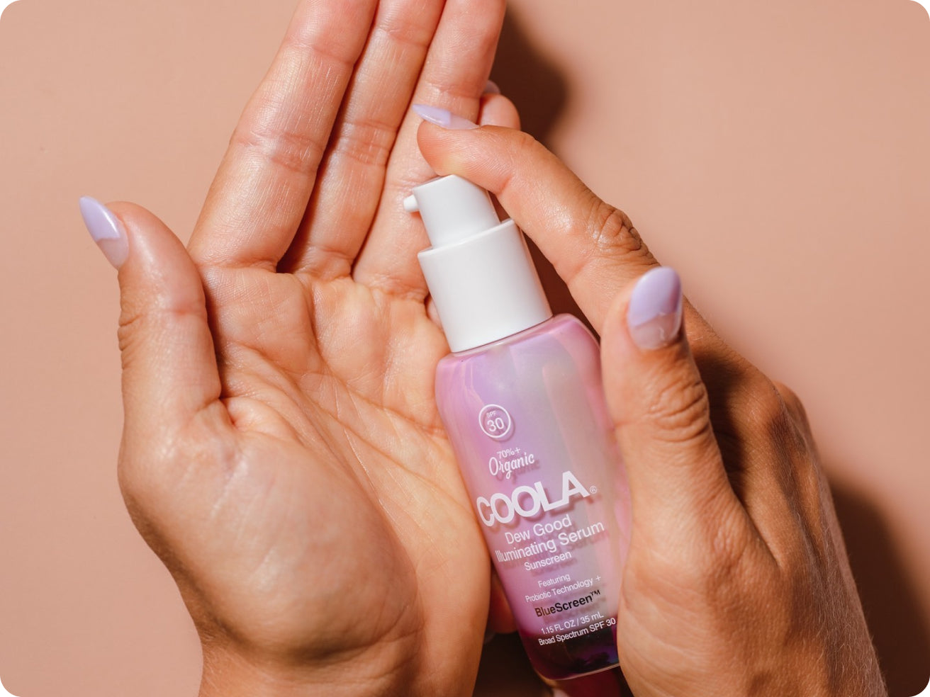 A hand with light purple nail polish dispenses COOLA Dew Good Illuminating Serum from a pump onto their palm against a peach-colored background.