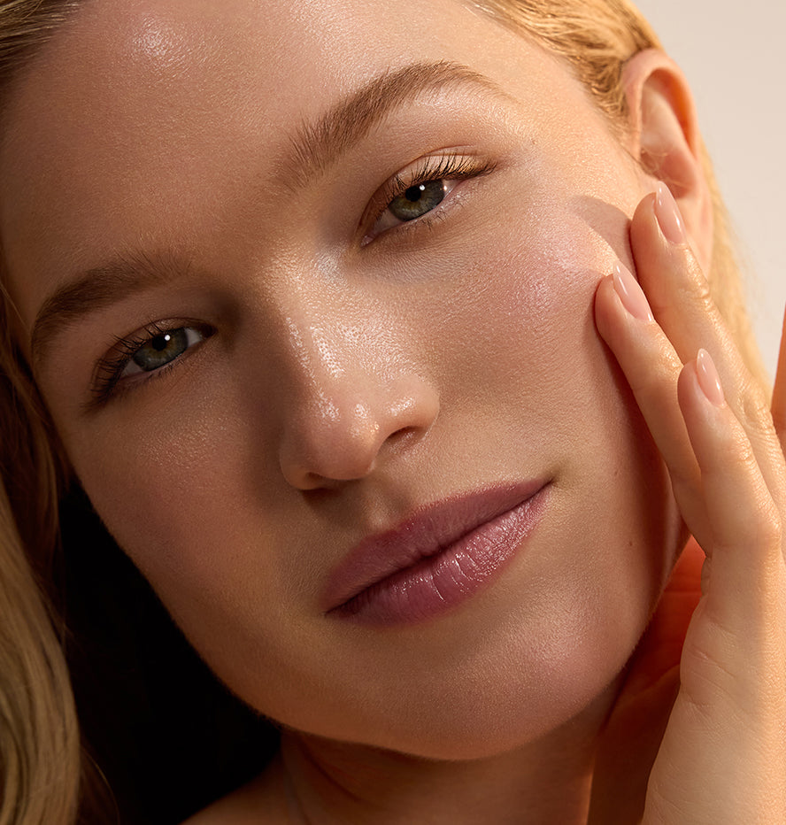 Close-up portrait of a woman with glowing dewy skin gently touching her cheek in warm natural light