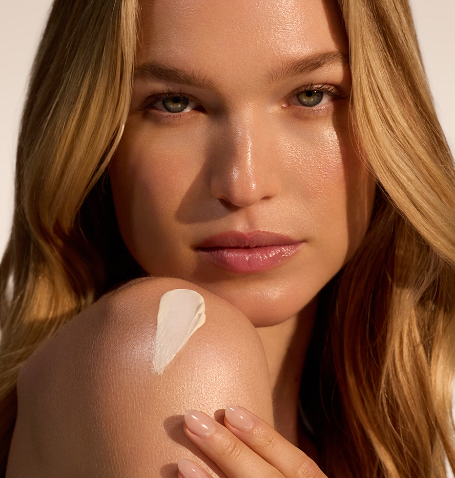 Close up of a woman showing a white sunscreen swatch on her shoulder with her hand resting nearby in warm sunlight